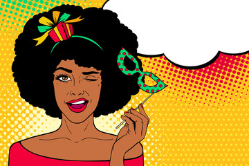 Wall Mural - Pop art face. Young sexy afro american woman with gift on her head and carnival mask in her hand smiles and winks with speech bubble. Vector illustration in retro comic style. Party invitation.