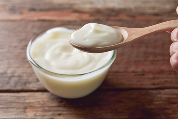 Yogurt in wood spoon on women hand and glass bowl on wooden tabl