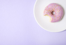 A Ring Doughnut With Pastel Pink Frosting And Sprinkles On A Purple Background, With A Bite Missing And Empty Space At Side