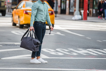 Elegant man walking on city street crosswalk wearing casual clothes with jeans ant t-shite and holding travel bag and sunglasses in hands