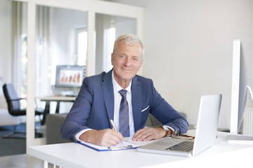 Senior professional man at work. Shot of an old financial businessman sitting at his workstation in front of computer and laptop while doing some paperwork.