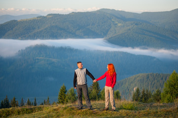 Couple holding hands and looking at each other standing on a hill against beautiful background with morning haze over the mountains.