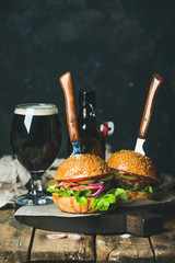 Wall Mural - Homemade beef burgers with crispy bacon and fresh vegetables on serving board with glass of dark beer on rustic wooden table, dark blue plywood wall as background, copy space, vertical composition