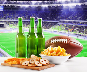 Wall Mural - Beer with snack and ball on table against football field background. Sport and entertainment concept.