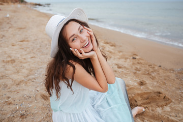 Wall Mural - Cheerful lovely young woman sitting and smiling on the beach