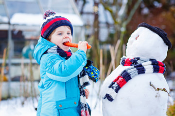 Poster - Funny kid boy in colorful clothes making a snowman, outdoors