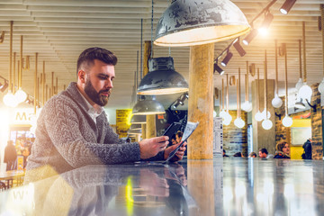 Sticker - Side view of young bearded businessman in gray cardigan sits at table in cafe,reading newspaper, using smart phone.From ceiling hang lamp.Man checking email,browsing internet,reading news, chatting.