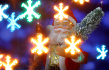 Close Up Santa Claus Toy Brings Christmas Tree At Blue Snowy Night Bokeh  Background And Blurred Lights Snowflakes Foreground. Big Copyspace Concept New Year`s Market Banner, Poster.