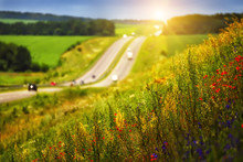 On The Slope Along The Road Bright Colors Poppies And Other Wild Flowers. In The Background In Soft Focus Road Fleeing Away To The Horizon. The Sun Low On The Horizon Illuminates 