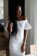 Adorable Attractive Femine With Black Skin Standing Near The Table. Girls Is Wearing Elegant White Dress With Sexy Bare Shoulders. She Looking At The Window And Put Her Hand On Hair.