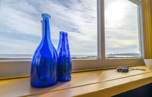 Blue Bottles In A Window Beside The Sea, Newfoundland Canada.
Wide Angle View, Blue Glass Bottles Decorate A Shelf At A Window With A View To The Atlantic In Grand Bank Newfoundland.