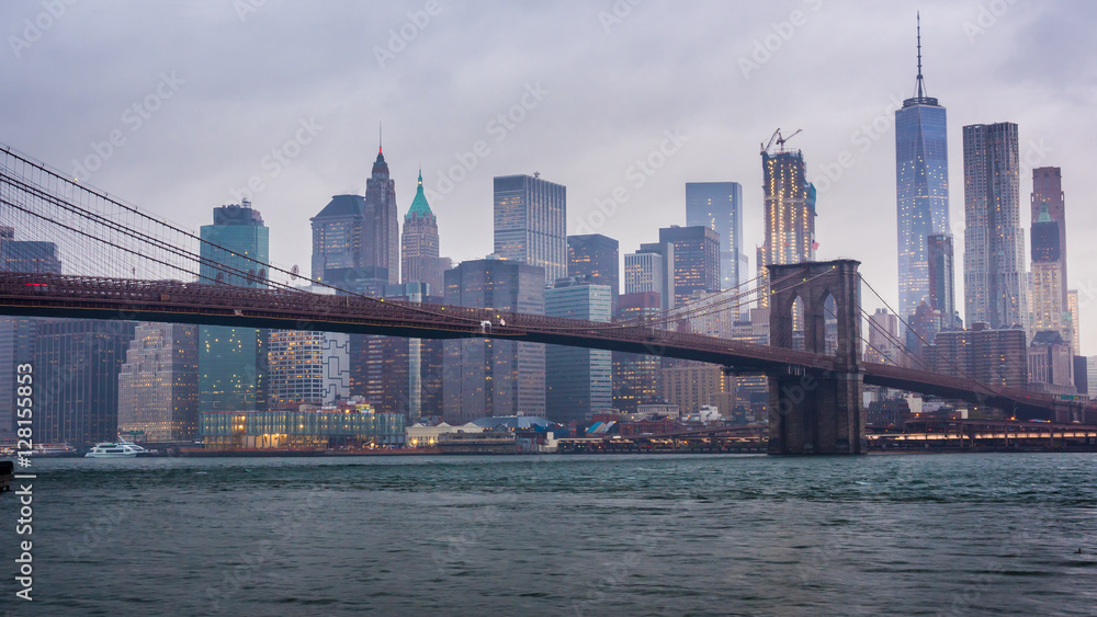 Timelapse day to night. Rainy Manhattan and the Brooklyn Bridge. The tops of the skyscrapers in clouds drown. Night comes to the business district of New York City - obrazy, fototapety, plakaty 