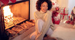© Dash - Woman seated by fireplace and holiday setting and wearing warm woolen sweater and white leggings. She has Christmas tree behind her back.