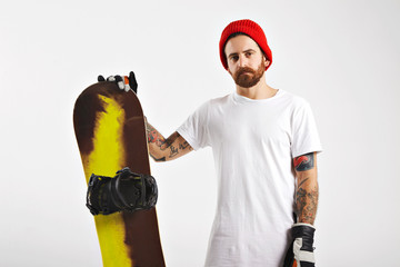 Wall Mural - Tattooed and bearded young man in plain white cotton t-shirt, red beanie and leather snowboard glasses holding a snowboard isolated on white