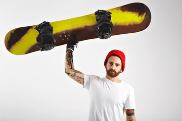 Wall Mural - Serious athletic young man with tattoos and beard raising his snowboard with one hand isolated on white