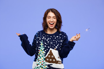 Young cheerful beautiful brunette girl in cosy knited sweater smiling holding bengal lights over blue background. 