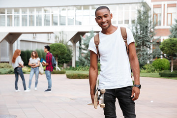 Poster - Cheerful african man student with backpack holding skateboard outdoors