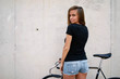 A brunette girl with long hair wearing a blank black t-shirt is standing with the back to the camera on a gray wall background on a street. Empty space for text or design.