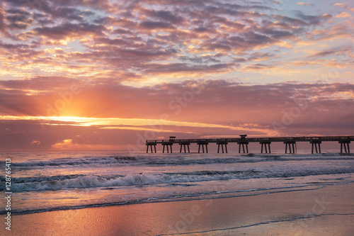 Fotografie, Obraz Sun rising over horizon and pier, beach illuminated with sunlight, beautiful sky reflected on the beach