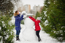 Mother With Daughter Of Younger School Age Build A Snowman In The Park.