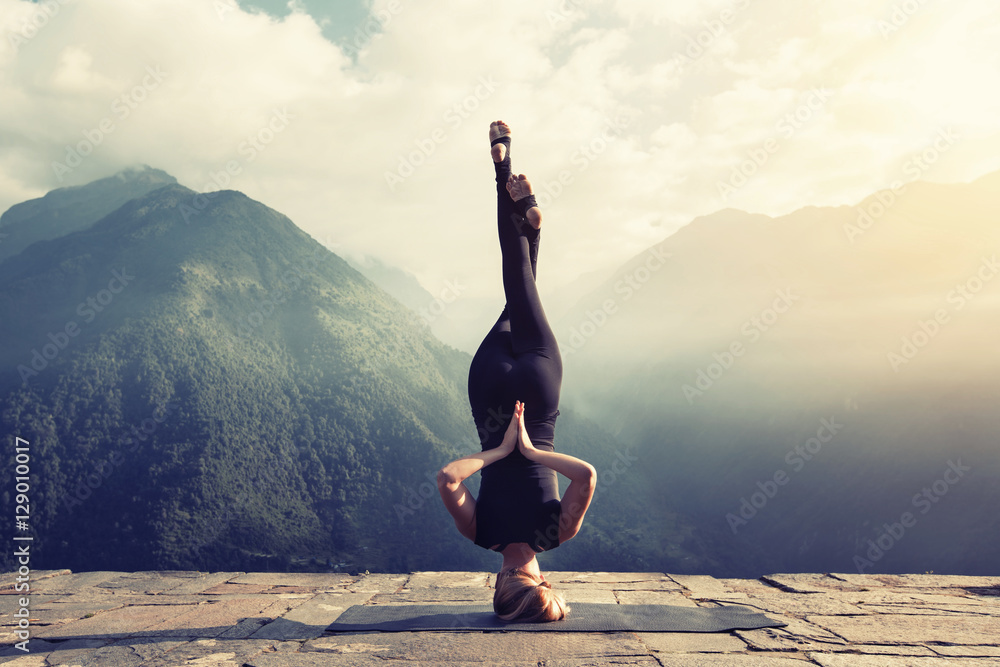 Young woman doing complex Yoga exercise headstand with Namaste asana. Amazing Yoga landscape in beautiful mountains - obrazy, fototapety, plakaty 