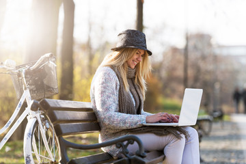 Blonde student girl with hat on park bench working on laptop