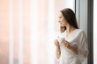 © fizkes - Young smiling woman in lace night gown relaxing at cozy home, feeling happy, resting in the morning, looking through the window with a cup of drink, challenge of a new day. Copy space