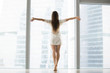 © fizkes - Young model in modern high rise building room with full length windows, outstretching arms, welcoming new day, dancing bare foot in morning, bought new flat. Feeling happy, alive, dreaming. Rear view