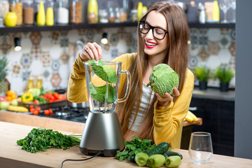 Young smiling woman making smoothie with fresh greens in the blender in kitchen at home. Healthy vegetarian smoothie for weight loss and detox