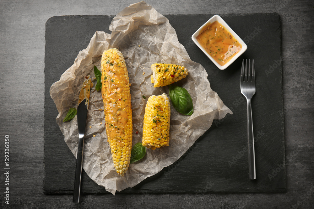 Slate plate with tasty grilled corncobs, basil, sauce, knife and fork on grey background