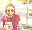 Young handsome man in sunglasses and headphones listening to music in a park outdoor sitting on a bench - Concept about people and technology