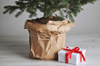 Pot with a Christmas tree wrapped in paper next to a cute present in a white box with a red ribbon on wooden floor next to a white wall.