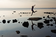 Silhouette Of A Young Girl Soaring In A Jump On The Beach At Sunrise With Reflection In Water