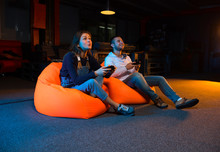 Two Young Gamer Sitting On Poufs And Playing Video Games Togethe