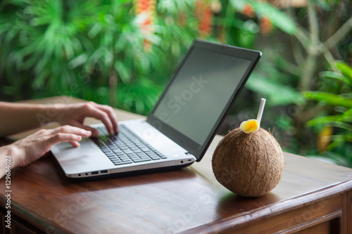 Fotografie, Obraz Tropical freelance: coconut in focus and worker's hands typing on keyboard of laptop