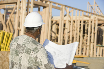 Senior man wearing hardhat with blueprint in front of incomplete house