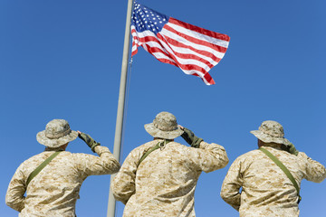 rear view of three soldiers saluting an American flag against blue sky