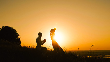 Silhouette of Man Getting Down on his Knee and Proposing to Woman high hill - Couple Gets Engaged at Sunset - Putting Ring Girl's Finger