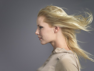 Closeup side view of a young woman with blond hair blowing in wind against gray background