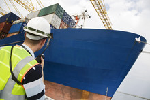 Rear View Of A Man In Hard Hat Using Walkie Talkie At Container Terminal