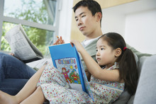 Daughter With Picture Book Sitting Beside Father On Sofa With Newspaper