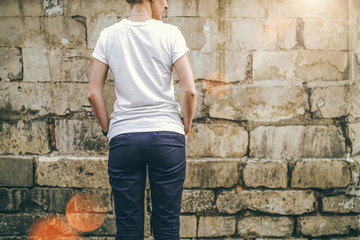 Wall Mural - Summer day. Back view. Young woman in a white T-shirt and blue pants standing on a background of a stone wall with his hands in his pockets. Cropped image. Mock up.