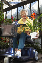 Portrait Of Disabled Senior Woman On Motor Scooter Holding Potted Plant At Botanical Garden