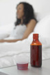 Cough syrup bottle and cup with sick woman in background