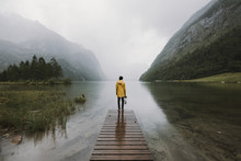 Young Adult Male Wearing Yellow Jacket Standing On A Footbridge At A Foggy Mountain Lake With Forest 