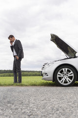 Wall Mural - Side view of young businessman using cell phone by broken car at countryside