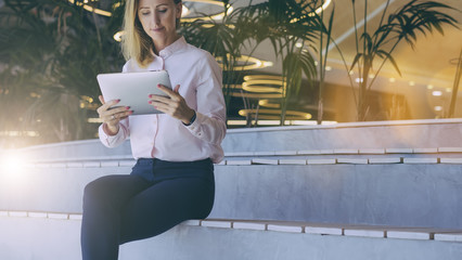 Wall Mural - Front view. Young businesswoman with blonde hair in pink shirt sitting in room with modern interior and uses tablet computer. Girl holding digital tablet and looking at its screen. Woman uses gadget.