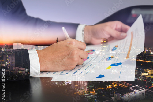 Fotografia, Obraz Double exposure of businessman hand hold a pen write, review document, cityscape, urban and street in the night as review and working Concept
