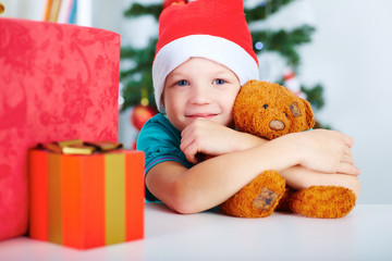 Boy with Christmas gifts and Teddy bear in hands, Christmas tree in the background. Christmas holiday concept.