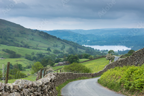 Fotografie, Obraz Lake District, England - May 30, 2012: Rural road with stone walls on the side meanders through the landscape